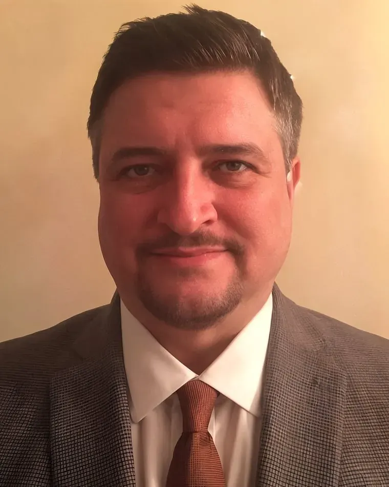 Mark Holland, smiling, in a gray check jacket, white shirt, and rust tie